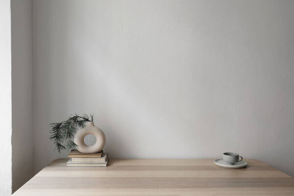 Modern vase on old books with pine tree branches.Cup of coffee on beige table.Empty white wall background in soft light. Minimal Christmas home decor, modern scandinavian interior. Winter design.