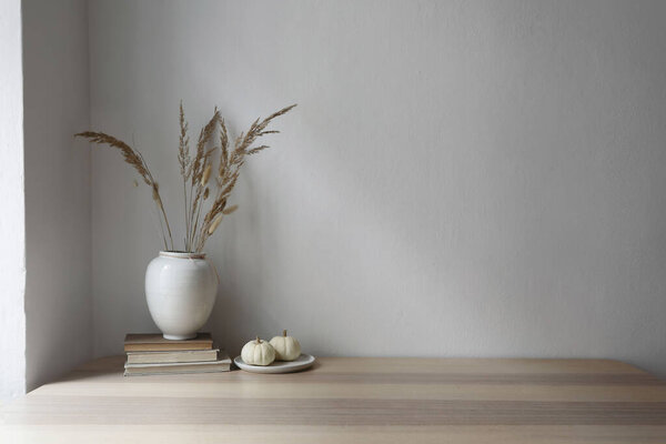Elegant moody Scandinavian interior. Vase with dry grass bouquet, old books and little white pumpkins. Empty wall background. Modern living room, home office, minimal autumn decor in cozy apartment.
