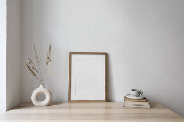 Elegant neutral poster mockup. Wooden picture frame mockup on beige table. Modern boho vase with dry grass, old books. Cup of coffee. White wall background, trendy Scandinavian interior, home office.
