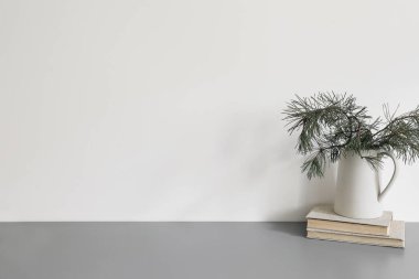 Elegant Christmas interior, home office. Ceramic vase, jug with green pine tree branches on old books. Winter home decor. Empty white wall background mockup. Grey table, desk, no people. clipart