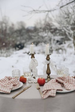 İki kadeh şarap, mum ve kareli peçeteli açık hava Noel sofrası. Kış düğünü, Sevgililer Günü, doğum günü partisi konsepti. Bulanık manzara, karlı bahçe. Dikey