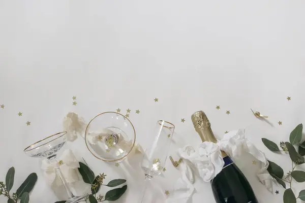 New year party, celebration banner. Bottle of champagne wine, drinking glasses, bows. Eucalyptus tree branches isolated on white table background. Golden star confetti decor, birthday flat lay, top