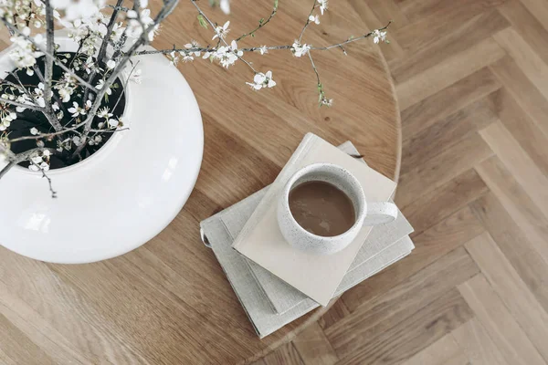 Spring breakfast scene. Cup of coffee on books. Round oak coffee table. Blurred parquet floor. Defocused blossoming cherry plum tree branches in vase. Top view, Easter still life, no people.