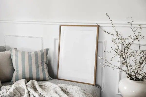 Modern spring scandinavian living room interior. Closeup of wooden picture frame, poster mockup. Sofa with linen beigecushions. Blurred cherry plum blossoms in vase. Selective focus. Elegant minimal