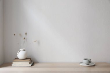 Modern iç mekan. Beyaz vazo ve kır tavşanı kuyruğu otu. Bir fincan kahve ve ahşap masada eski kitaplar. Boş bej duvar arkaplanı. Tarafsız model, İskandinav evi, oturma odası tasarımı.