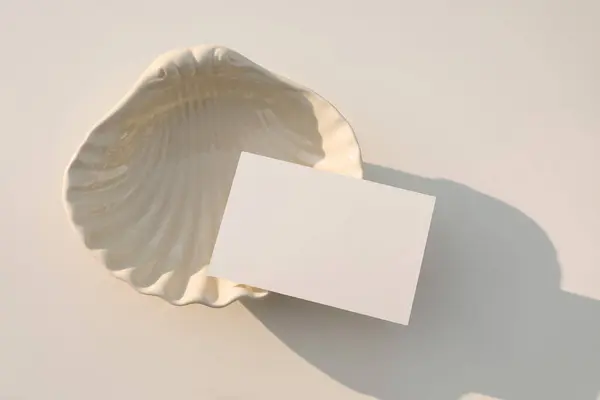 Summer neutral wedding stationery. Blank greeting, RSVP or business card mockup in sunlight. Invitation, template. Beige sea shell plate. White table background. Elegant composition, flatlay, top view