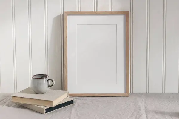 Blank vertical wooden picture frame mock up on linen table cloth. Modern interior. Cup of coffee on pile of vintage books. White wall background. Neutral Scandinavian home, poster display template.
