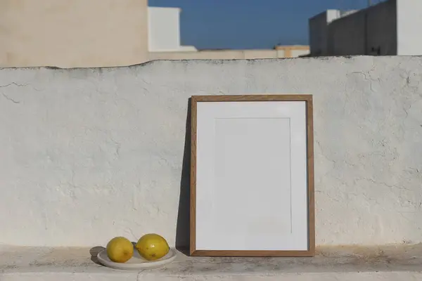 Blank vertical wooden frame picture mock up with fresh lemons fruit. White old textured white wall in sunlight. Summer display background for art, posters. Blurred blue sky, houses. Roof tops.