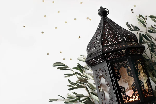 Glowing dark Moroccan, Arabic lantern and olive tree twigs isolated on white table background. Golden star confetti. Festive still life, banner. Muslim holiday Ramadan Kareem, flat lay, top view.