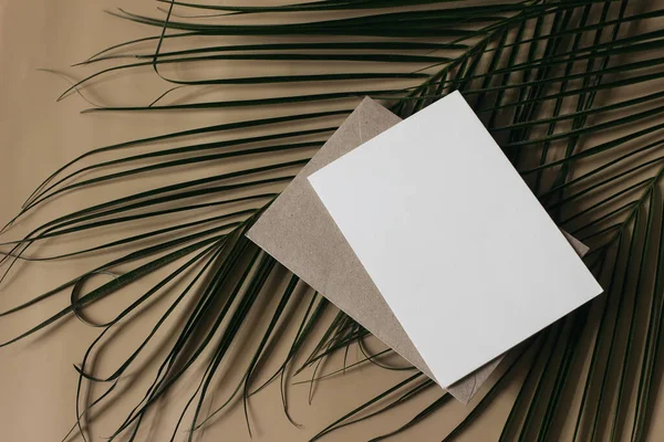 Summer tropical wedding stationery still life scene. Green date palm leaves. Blank greeting card, invitation mockup scene. Craft envelope. Beige table background. Flat lay, top view. No people.