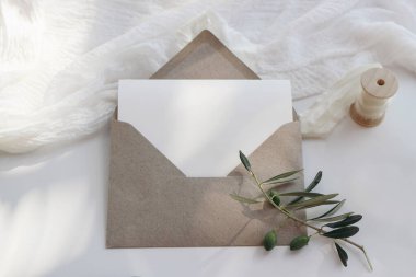 Yaz kırtasiyesi hala hayat konsepti. Boş tebrik kartı, açık el işi zarflı davetiye modeli. Zeytin dalları, beyaz masa arkasında muslin tekstili. Düğün davetiyeleri, üst manzara.