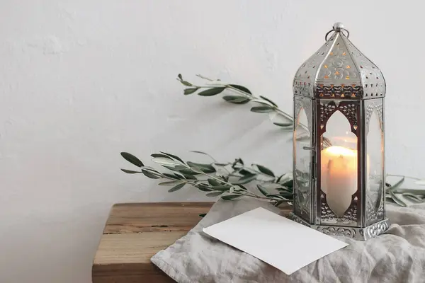 Ramadan Kareem holiday still life. Ornamental silver Moroccan lantern with olive tree branches. Blank greeting card, invitation mockup on wooden table, bench, blurred white wall background.