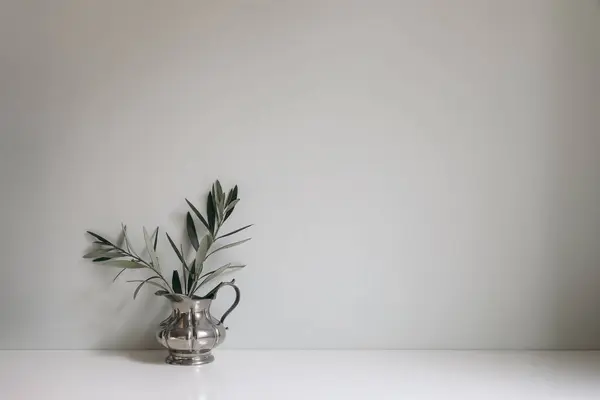 Olive tree branches, twigs in silver jug vase on white table. Empty mint green wall mockup, background. Working space, home office decor, vintage Mediterranean summer interior still life.