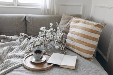 Modern bahar İskandinav oturma odasının iç kısmı. Keten sarısı çizgili yastıklı kanepe ve bir fincan kahve. Vazoda kiraz çiçekleri var. Boş bir kitap, günlük ya da not defteri, şık bir ofis dekorasyonu..