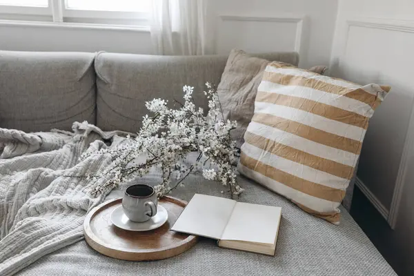 Modern spring scandinavian living room interior. Sofa with linen yellow striped cushions and cup of coffee. Cherry plum blossoms in vase. Open blank book, diary or notepad, elegant home office decor.