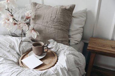 Yatak konseptinde kahvaltı. Tahta tepside bir fincan kahve. Boş tebrik kartı, davetiye maketi. Yatak odası manzaralı. Bulanık bej keten yastıklar, battaniye. Vazodaki çiçek açan manolya dalları, üst görünüm.