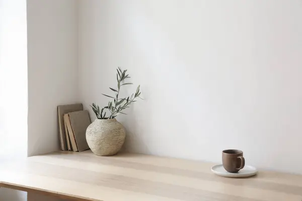 Breakfast, scandi indoor interior still life. Minimal home design concept. Beige ceramic vase with olive tree branches. Cup of coffee, tea on wooden table, desk with old books. Empty beige wall mockup