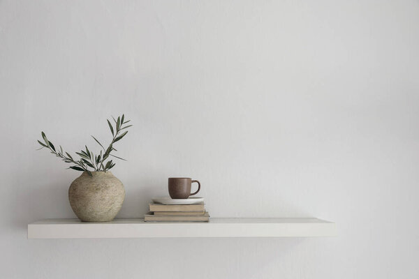 Elegant home still life. Floating shelf. Textured vase with green olive tree branches and old books. Cup of coffee, tea. Modern Mediterranean appartement. White wall background, interior indoor mockup