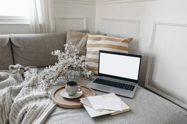 Spring scandinavian living room interior. Sofa with linen yellow striped cushions. Cup of coffee. Cherry plum blossoms in vase. Laptop mockup with blank screen and greeting cards, open book, home