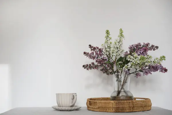 カップのコーヒー ウィッカートレイで静けさの瞬間 花の花束 ガラス花瓶に紫と白のライラックの枝が咲きました 春の朝食シーン テーブル 白い壁の背景 選択的 — ストック写真