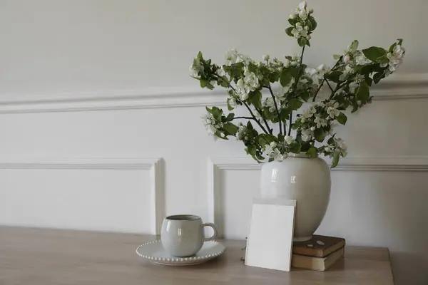 春の朝食はまだ生きている 空の挨拶カード 招待状のモックアップ リンゴの木の枝が咲いている白い陶磁器の花瓶 コーヒーのカップ 木のテーブルの紅茶 古い本 スキャンディの家の内部 — ストック写真