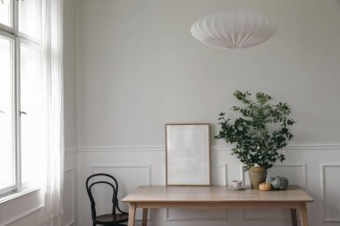 Modern İskandinav oturma odasının iç kısmı. Ahşap poster, ofis masasında resim çerçevesi modeli vazoda yeşil ağaç dalları, balkabakları, bir fincan kahve. Pencerenin yanındaki eski sandalye. Tarz ev dekoru.