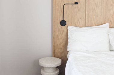 Büyük, boş, çift kişilik yataklı İskandinav tarzı yatak odası. Beyaz çarşaflar, yastıklar. Modern komodin, siyah duvar lambası. Rahat bir uyku. Meşe ağacından yatak başlığı. Gri duvar arkaplanı, otel konaklama