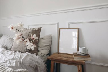 Blank wooden picture frame mockup on old book. Wooden night stand with fluted glass vase. Blooming magnolia tree branches. Scandinavian interior. Elegant bedroom, white wall background, stucco decor. clipart