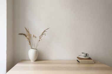 Zarif nötr iç modelleme. Kuru çimenli vazo, eski kitaplar, ahşap masada bir fincan kahve. Beyaz duvar arka planı. Modern İskandinav ev ofisi, apartman dekorasyonu, boş kopya alanı.