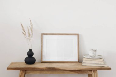 Modern siyah vazo, kuru çimen çiçekleri eski ahşap masa, bank. Bir fincan kahve, kitaplar. Yatay boş çerçeve modeli. Sanat, poster gösterisi. Modern iç mekan, beyaz duvar arkaplanı.