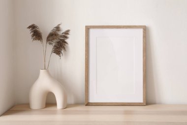 Modern seramik vazoda kuru ot, kamış çiçeği buketi. Boş çerçeve, ahşap masada poster modeli. Beyaz duvar arka planı. Minimalist İskandinav tarzı, zarif bir iç mekan. Oturma odası..