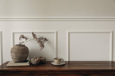 Zarif büfe sonbahar düzenlemesi. Minimalist aksanlı şık ahşap büfe dolabı. Kil vazo, kuru eğrelti otu yaprağı, balkabağı, bir fincan kahve. Modern iç tasarım trendleri. Beyaz duvar arkaplanı.