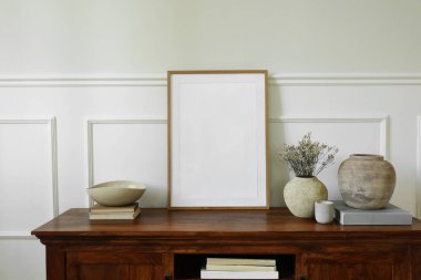 Zarif büfe düzenlemesi. Mhogany büfe dolabı. Boş dikey ahşap resim poster çerçevesi gibi. Kil vazolar, kuru çiçekler, kitaplar, mum. Modern iç tasarım. Beyaz duvar arkaplanı.