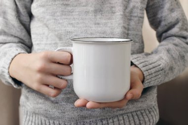 Eller yakın, parmaklar beyaz çay bardağını tutuyor. Boş cearmik kupa maketi. Bulanık arkaplan, seçici odaklanma. Kahve zamanı. Kış durgun, gri örgü kazaklı çocuk. Noel içeceği.