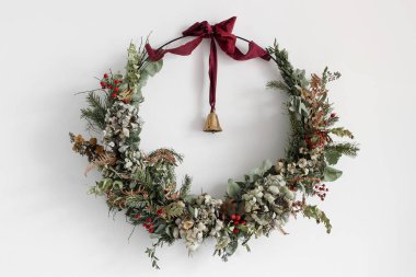 Köknar ağacı dalları, kırmızı böğürtlenler ve beyaz duvarda asılı kuru çiçeklerden oluşan harika bir çelenk. Kestane rengi kurdeleli altın çan. Minimum estetik kış tatili ev dekorasyonu, kopya alanı