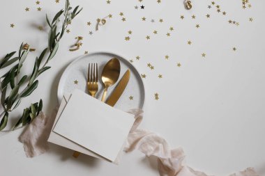 Happy New Year holiday party celebration. Olive tree branches isolated on white table background. Golden star confetti. Golden cutlery, silk ribbon on plate. Blank greeting card mockup. Festive flat lay, top clipart