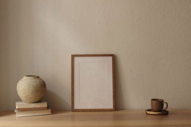 Zarif asgari ev ofisi hala hayat dolu. Masada boş ahşap çerçeve poster modeli var. Scandi iç dekorasyon. Bir fincan çay, kahve, eski kitaplar. Dokulu vazo. Bej duvar arkaplanı, boş kopya alanı.