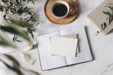 Zarif yaz kahvaltısı natürmort, kırtasiye. Boş tebrik kartı, davetiye maketi, açık günlük ve zeytin ağacı dalları, beyaz mermer masa arka planı. Bir fincan kahve. Bulanık yapraklar. Düz yatıyordu. Üst