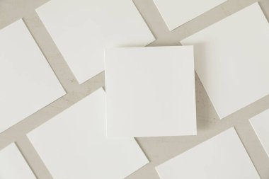 Pek çok boş beyaz kare tebrik kartı modeli yumuşak gölgeli bej desenli ahşap masa zemininde çapraz olarak yatıyor. Bir kart üstte. Düz konum, üst görünüm, kırtasiye bileşimi.