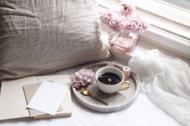 Bahar natürmort. Boş tebrik kartı davetiyesi maketi. Mermer tepside bir fincan kahve, eski kitaplar. Kadınsı bir fotoğraf. Çiçek kompozisyonu. Pembe sakura, kiraz ağacı çiçekleri. Pencerenin yanındaki tablo