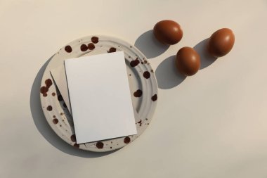 Paskalya 'da hayat. Kahverengi benekli seramik tabakta boş tebrik kartı, davetiye. Bej masa arkaplanındaki tavuk yumurtaları güneş ışığı, uzun gölgeler. Bahar zamanı tatil evi dekorasyonu. Düz yatıyordu. Üst görünüm