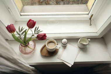 Sıcak Paskalya, bahar durgun hayat sahnesi. Bir fincan kahve, ahşap tabak, haşlanmış yumurta, vazo dolusu çiçek, pencere pervazı. Laleli çiçek kompozisyonu, zeytin dalları. Boş tebrik kartı davetiyesi maketi. Üst