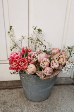 Kadınsı bahar Paskalyası. Pembe lale çiçekleri, çiçek açan kiraz ağacı dalları metal kovada. Bulanık ahşap ön kapı arka planı. Çiçekli düğün süsü, dikey. Üst görünüm.