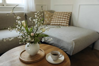 Huysuz bahar iç mekanı hala hayat dolu. Bir fincan kahve. Yuvarlak ahşap masa. Laleler vazoda kiraz ağacı dalları açıyor. Penceresinde yastıklar olan rahat keten kanepe. Zarif oturma odası manzarası, ev