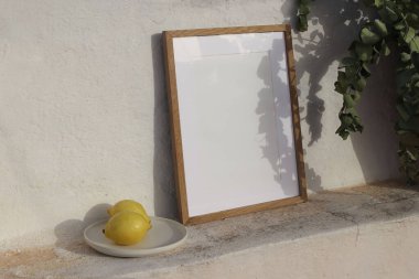 Minimalist yaz durgun hayat sahnesi. Beyaz desenli bir duvara yaslanmış boş ahşap çerçeve modeli. Seramik tabak, iki taze limon, güneş ışığıyla yıkanmış. Yapraklı gölgeler, okalipsiz dallar