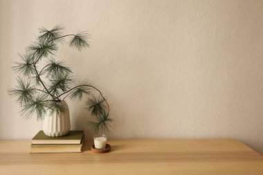 Minimalist Noel iç tasarımı, natürmort. Yığılmış kitapların üzerinde çam ağacı dalları olan beyaz vazo, ahşap masa üzerinde cam tutacağında küçük mum bej duvar arka planında, Varm kışlık ev ofisi.