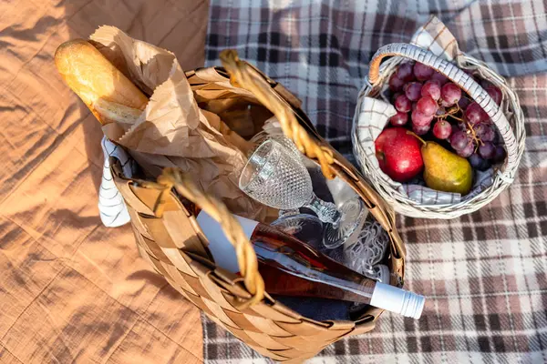 Şarap ve ekmekle hasır sepetin yanı sıra bir sepet taze meyvenin olduğu bir piknik düzeneği, güneşli bir açık hava ortamında kareli bir battaniyeye yerleştirilmiş, rahatlama ve rahatlama çağrıştıran