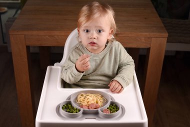 Yüksek sandalyede oturan bir çocuk spagetti ve bezelye yiyor. Bebek menüsü, çocuk menüsü, eğlenceli keşifler, günlük çocukluk rutinleri, kendini besleme, bebek sütten kesilmeye yol açtı..