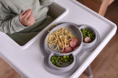 Yüksek sandalyede oturan bir çocuk spagetti ve bezelye yiyor. Bebek menüsü, çocuk menüsü, eğlenceli keşifler, günlük çocukluk rutinleri, kendini besleme, bebek sütten kesilmeye yol açtı..