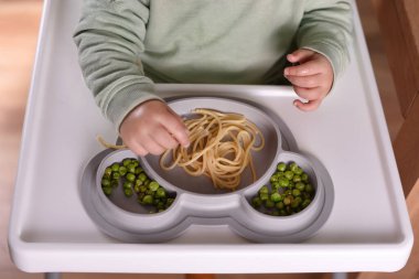 Yüksek sandalyede oturan bir çocuk spagetti ve bezelye yiyor. Bebek menüsü, çocuk menüsü, eğlenceli keşifler, günlük çocukluk rutinleri, kendini besleme, bebek sütten kesilmeye yol açtı..